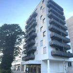 Weißes 9 stöckiges Haus mit schwarzen Balkonen und Glaseingang, junger Mann steht vor einem Roller und schaut in sein Rucksack. Hausansicht von der Straße in der Ostparkstr. 45, 60385 Frankfurt am Main